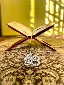 An open Quran on a wooden stand with prayer beads on a decorative carpet, reflecting Islamic spirituality.